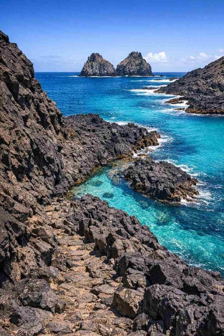 A dramatic view from the Baia dos Porcos guide showing black volcanic rocks against turquoise water and the Morro Dois Irmãos.