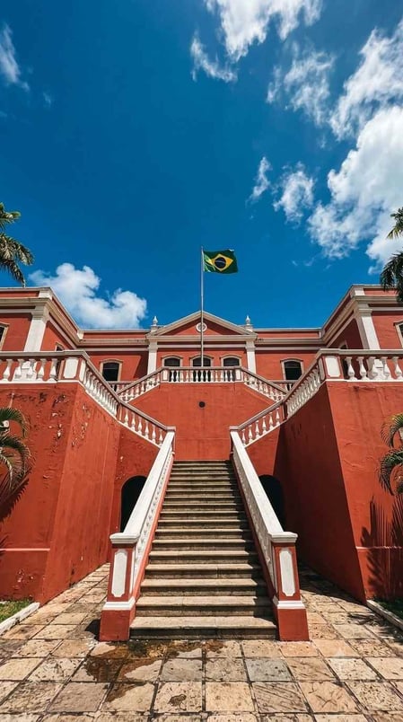 The grand red façade of the São Miguel Palace in Fernando de Noronha, Brazil, with text overlay asking "A Palace Built on Prison Ruins?"