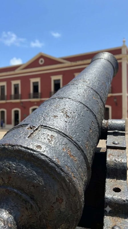 Un primo piano di un cannone storico a Fernando de Noronha, con il Palazzo São Miguel rosso sullo sfondo.