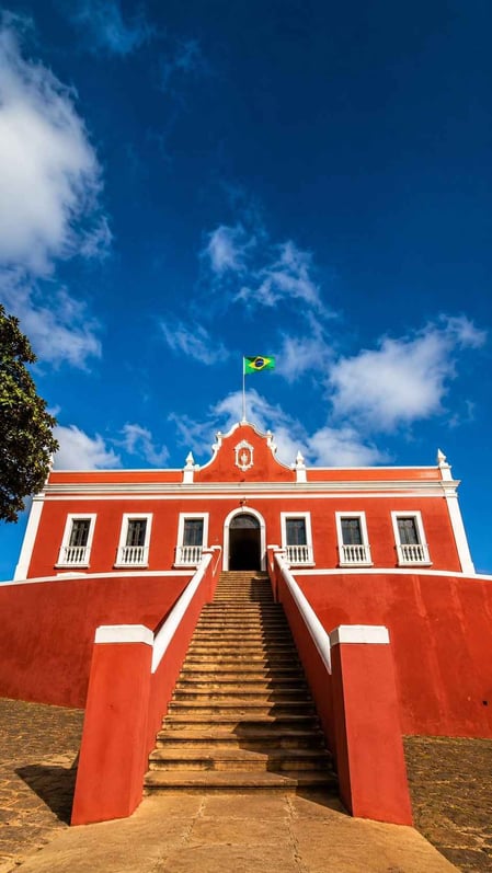 La maestosa facciata rossa del Palazzo São Miguel a Fernando de Noronha, in Brasile, con una didascalia che recita: «Un palazzo costruito sulle rovine di una prigione?».