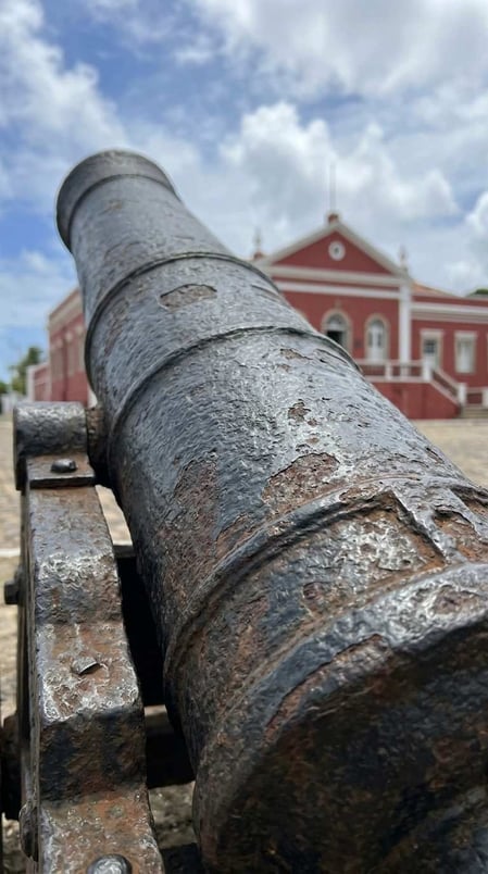 Um close-up de um canhão histórico em Fernando de Noronha, com o Palácio de São Miguel, de cor vermelha, ao fundo.