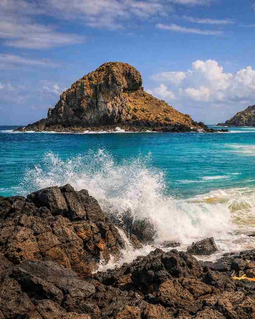A icônica rocha em forma de leão na Praia do Leão, em Fernando de Noronha, com ondas poderosas quebrando na areia dourada, conforme apresentado em nosso guia de viagem da Praia do Leão.